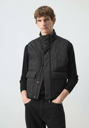 Hombre con chaleco negro acolchado con bolsillos delanteros sobre camisa negra de manga larga, de pie con las manos en los bolsillos frente a un fondo neutro.