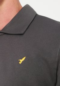 Polo noir avec un petit oiseau jaune brodé sur la poitrine gauche, zone visible du cou et du col.