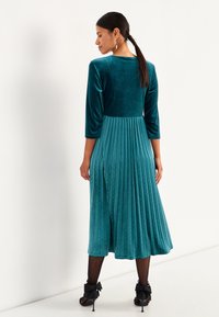 Türkisfarbendes Samtkleid mit drei Viertelärmeln, gefälteltem Rock und tailliertem Oberteil. Besitzt eine glatte Textur und einen subtilen Glanz.