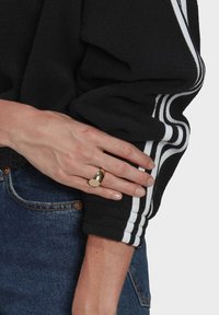 Kultainen metallisormus, jossa on pyöristetty yläosa, lepää kädellä. Kädellä on musta fleece-takki, jossa valkoiset raidat, ja siniset farkut.