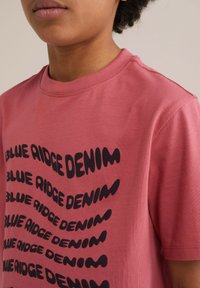 Ružičasta pamučna majica s kratkim rukavima, na kojoj se ponavlja crni natpis "BLUE RIDGE DENIM" u valovitom fontu. Klasični kroj s okruglim izrezom.