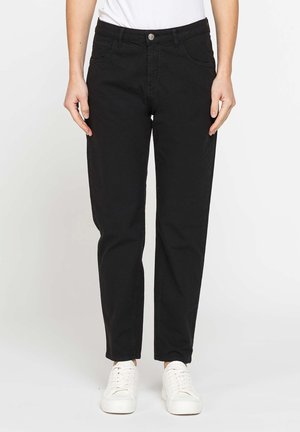 Persona che indossa jeans neri dritti, sneakers bianche e una maglietta bianca, in piedi davanti a uno sfondo bianco uniforme.