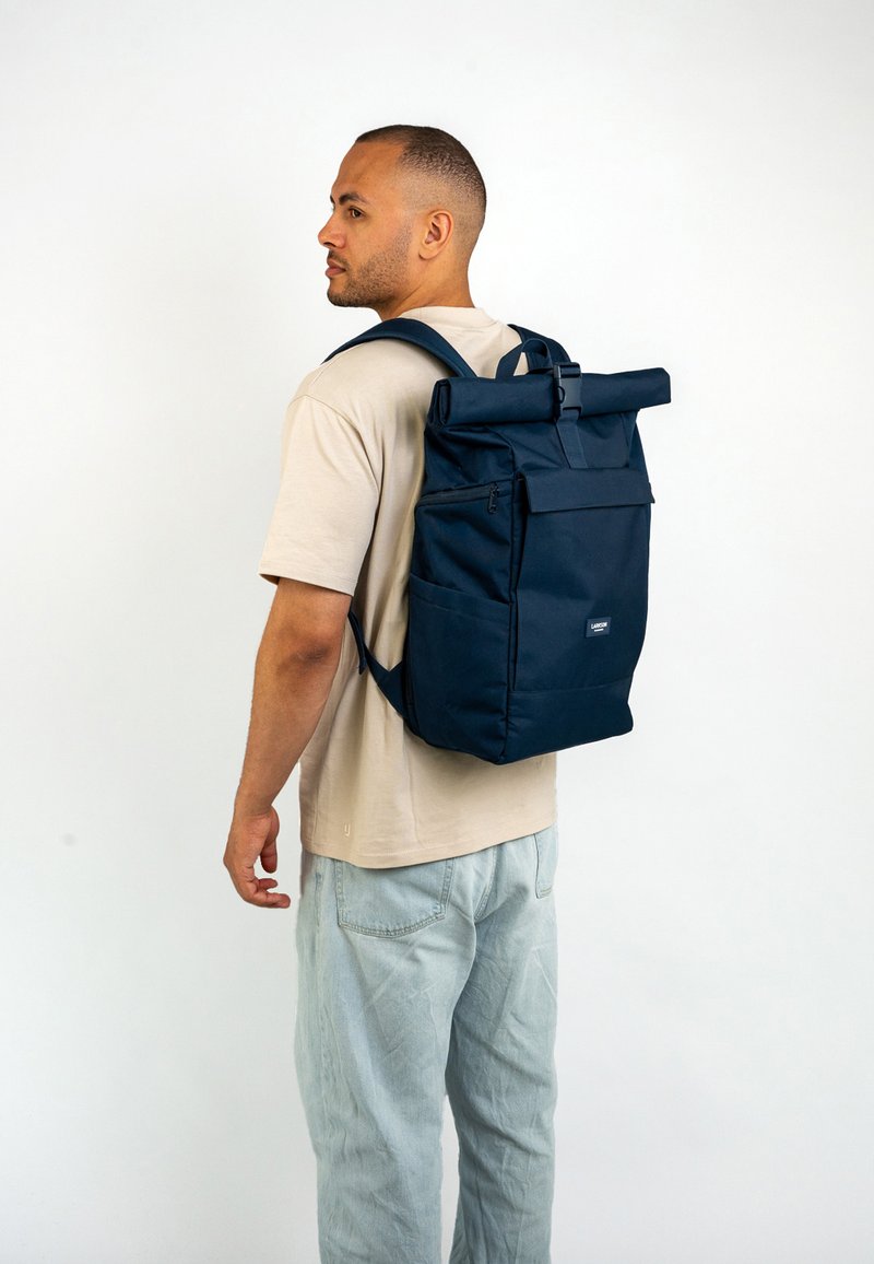 Sac à dos en toile marine à rabat, avec des bretelles ajustables, une poche avant et un tissu texturé. Modèle portant une chemise beige et un jean clair, de dos.