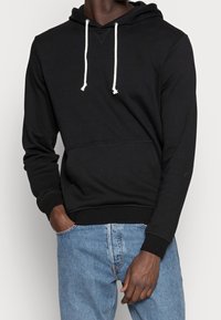 Hombre con sudadera negra con cordones blancos y jeans azules, con una mano en el bolsillo, frente a un fondo claro y liso.
