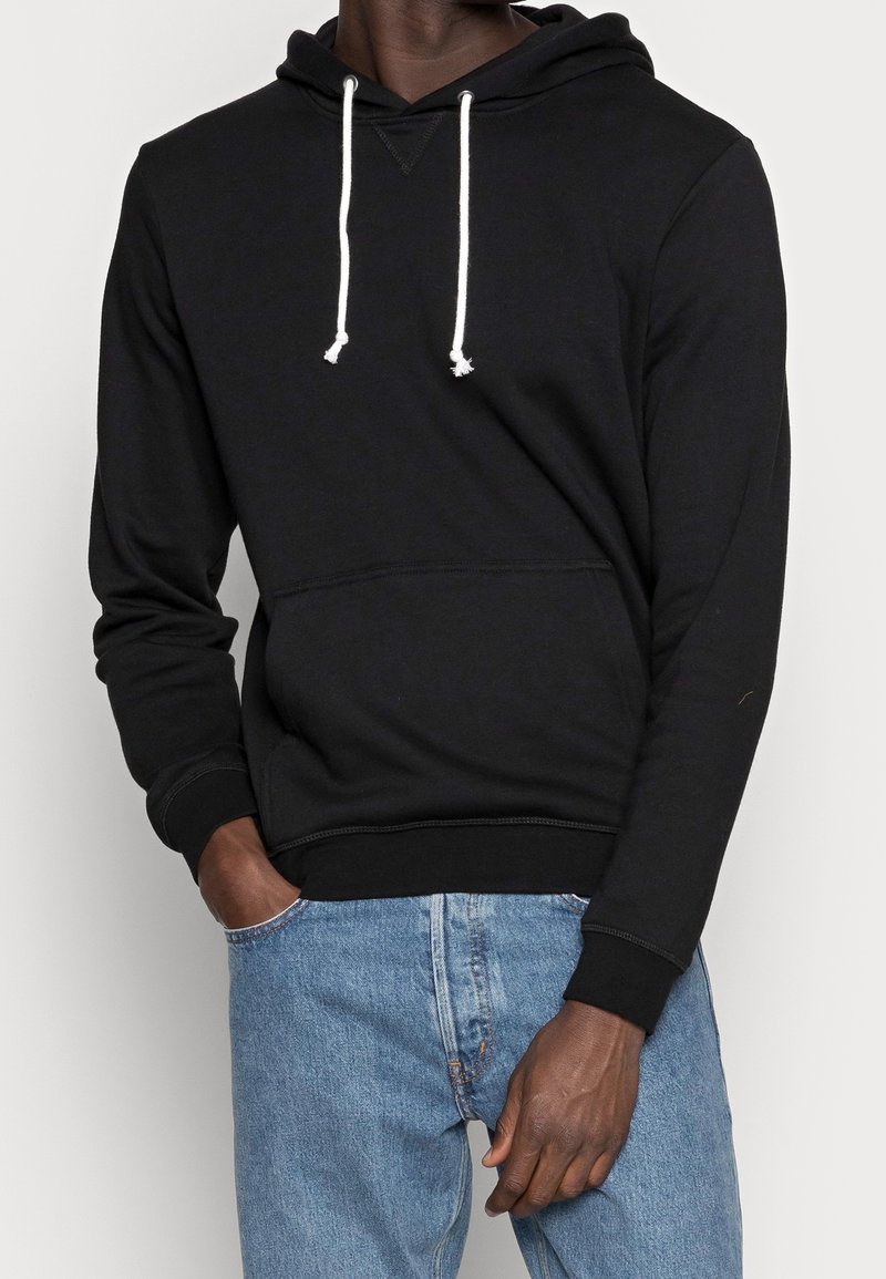 Hombre con sudadera negra con cordones blancos y jeans azules, con una mano en el bolsillo, frente a un fondo claro y liso.