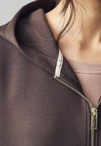 Gros plan d'une personne portant un sweat à capuche marron zippé par-dessus un haut côtelé rose clair, avec des mèches de cheveux lâches près du cou et la fermeture éclair partiellement fermée.