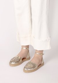 Zlaté espadrilky s kulatou špičkou, dekorativním výšivkovým designem, pletenou jutovou podešví a nastavitelným páskem na kotníku.