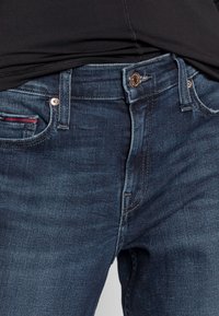 Jeans in denim blu scuro con vestibilità slim, dotati di un bottone in rame, due tasche frontali e cuciture di accento rosse sulla tasca sinistra.