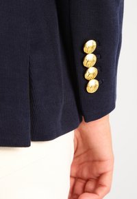 Manche de veste bleu marine avec quatre boutons dorés brillants près du poignet et une main reposant à côté du pantalon blanc.