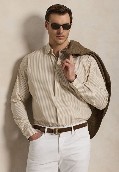 Man in bruin gestreept overhemd, witte broek, bruine riem, zonnebril, houdt een bruine jas over de schouder tegen een effen achtergrond.