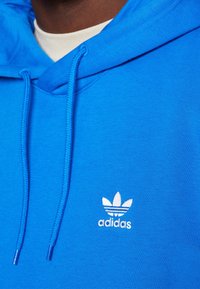 Modrá mikina s kapucí a stahovací šňůrkou, s bílým vyšívaným logem Adidas a detaily tří pruhů. Vyrobena z měkké, strukturované tkaniny.