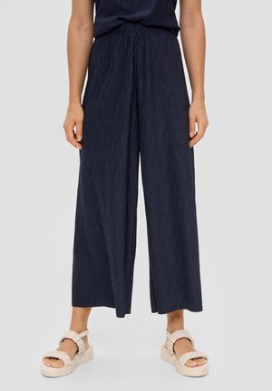 Vrouw draagt donkerblauwe wijde cropped broek en crèmekleurige gevlochten sandalen, staand tegen een effen achtergrond.