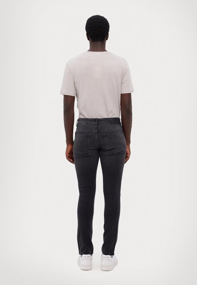 Jean skinny noir à taille haute, avec une texture lisse, deux poches arrière, et un discret logo sur la ceinture.