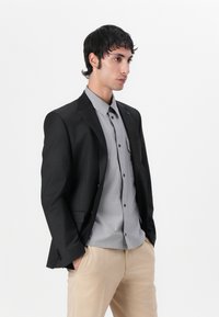 Blazer negro sobre una camisa gris, ambos ajustados para un look entallado. Pantalones beige. Texturas suaves, elementos de diseño sutiles y líneas limpias.
