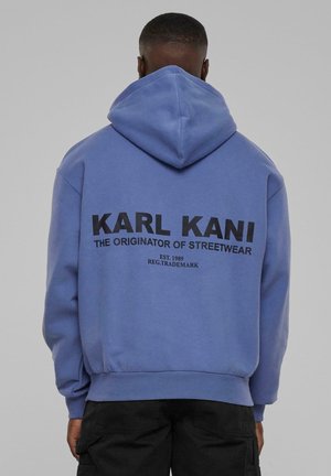 Mężczyzna w niebieskiej bluzie z kapturem z napisem „KARL KANI THE ORIGINATOR OF STREETWEAR” na plecach, czarne spodnie, stoi na jednolitym tle.