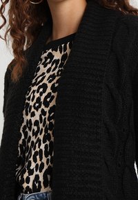 Czarny dzianinowy kardigan z fakturą splotu warkoczowego, noszony na topie w panterkę. Posiada szeroki kołnierz i luźny krój.