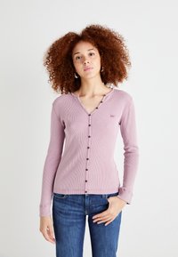 T-shirt à manches longues en lavande douce avec un col en V et des boutons foncés sur le devant. Dispose de poignets ajustés et d'un petit détail de logo.