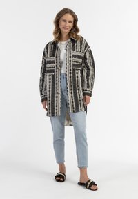 Raštuota juoda ir balta per didelė bliuzono marškiniai su ilgomis rankovėmis, derinama su šviesiai mėlynomis džinsomis ir juodais šlepetėmis, papuoštomis karoliukais.