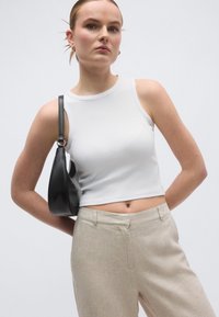 Λευκό κοντό τοπ χωρίς μανίκια συνδυασμένο με ανοιχτό μπεζ παντελόνι. Το μοντέλο κρατάει μια μαύρη δερμάτινη τσάντα με καμπυλωτό σχήμα και ιμάντα ώμου.