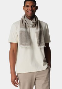 Beige Schal mit strukturiertem Finish, der weiße Streifen und fransige Kanten aufweist, um ein helles Hemd gewickelt, präsentiert an einem männlichen Modell.