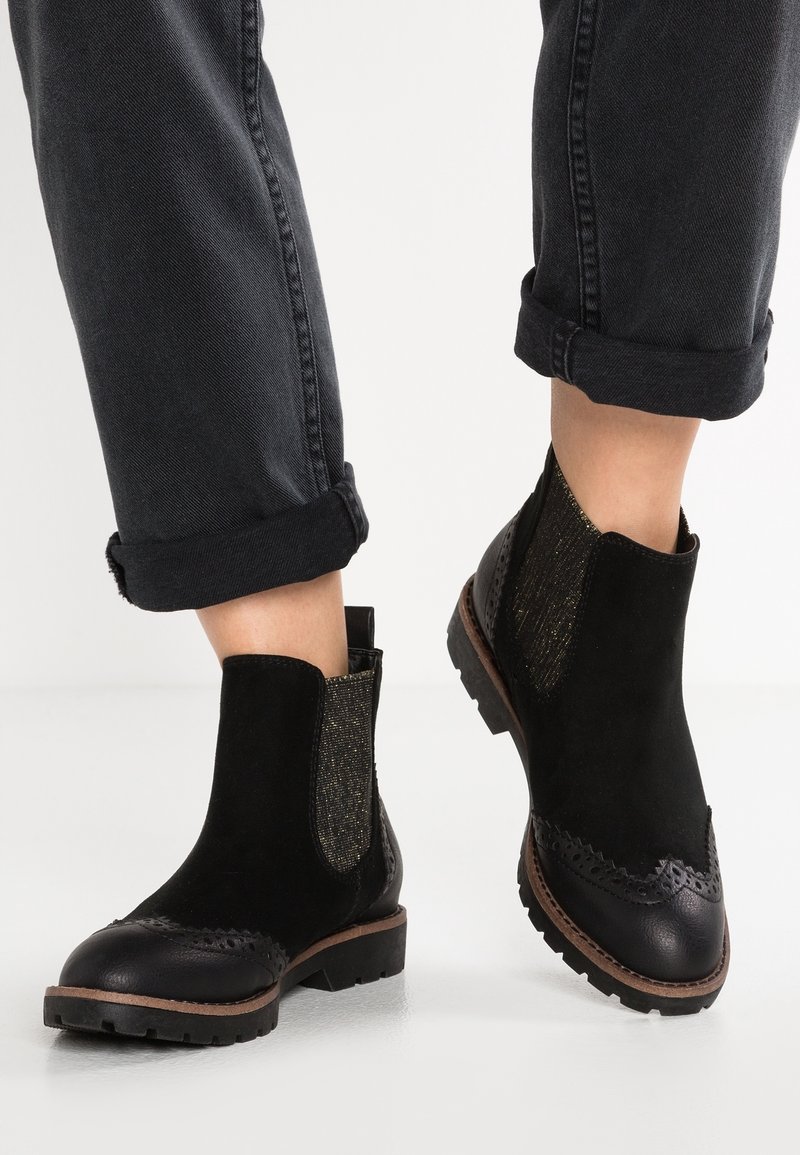 Bottes noires à chevilles avec tige en daim, panneaux latéraux élastiques et bout en cuir noir texturé. Détail brogue remarquable sur le bout renforcé.