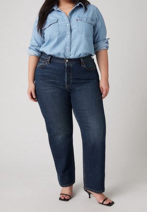 Vrouw draagt een lichtblauw denim overhemd en donkerblauwe hooggesloten spijkerbroek met zwarte open teen sandalen met hak.