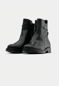 Une paire de bottes noires en cuir à cheville avec des talons bas, des zips latéraux et des sangles décoratives sur un fond gris clair uni.