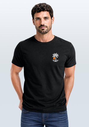 Mann mit dunklen Locken und Bart, der ein schwarzes T-Shirt mit kleinem weißem Palmbaum- und orangem Sonnenmotiv trägt, steht vor hellem Hintergrund.