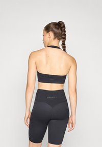 Črn športni crop top s halter dizajnom, v kombinaciji z visokim pasom črnimi kratkimi hlačami. Obe imata gladko teksturo in prikazujeta blagovno znamko "STRONGER".
