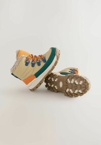 Bottines tan et vert avec doublure en fourrure, ornées d'accents orange et de œillets bleus. Semelle en caoutchouc chunky et texturée avec une adhérence à motif.