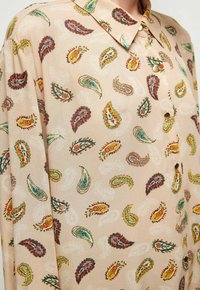 Blusa beige con un patrón paisley en rojo, azul y amarillo. Cuenta con un cuello y un cierre de botones. Tela suave y ligera.