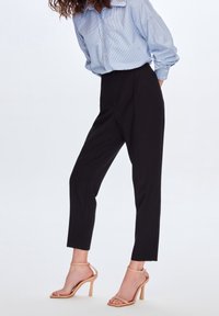 Femme portant un pantalon noir taille haute tapered, une chemise à rayures bleues à boutons, et des sandales à talons hauts à bride cheville couleur nude, se tenant sur un fond blanc.