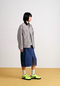 Grijze jas met een zwarte kraag, gedragen over een witte top. Spijkerrok met een split aan de voorkant, gecombineerd met neongele sneakers en witte sokken.