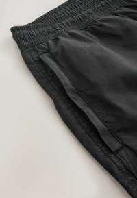Gros plan sur la ceinture élastique noire et la poche latérale zippée sur un pantalon en tissu léger, sur un fond neutre.
