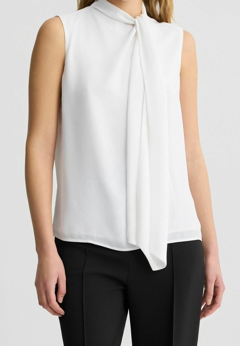 Blouse blanche sans manches avec un détail de col noué associée à un pantalon noir portée par une personne sur un fond uni.