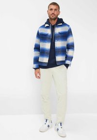 Giacca a quadri blu e bianca con due tasche sul petto, indossata sopra un hoodie blu navy. Pantaloni in corduroy crema e sneaker bianche con accenti blu.