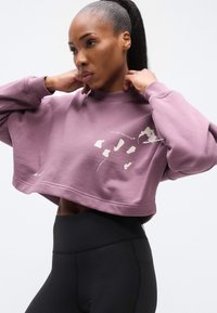 Μωβ κοντή φούτερ μπλούζα με ριμπ γιακά και φαρδιά μανίκια. Διακοσμημένη με απλό και γραφικό σχέδιο στο μπροστινό μέρος. Συνδυασμένη με μαύρα κολάν.