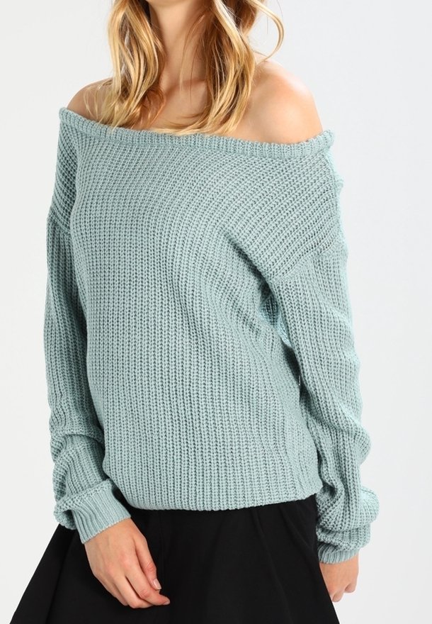 Femme portant un pull tricoté épais bleu clair à épaules dénudées, associé à une jupe noire, posant devant un fond uni.