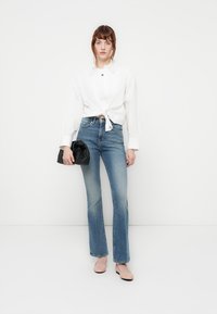 Camicia bianca a maniche lunghe legata in vita, jeans a campana blu e una pochette nera. Silhouette aderente con una morbida texture in denim e un leggero gradiente di colore.