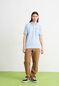 Světle modré polo tričko s kontrastními tmavými pruhy, spárované s béžovými kalhotami. Model stojí na zelené podlaze a má na sobě černé tenisky.