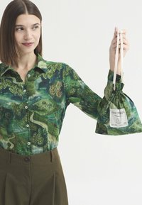 Femme portant une chemise verte à motifs et un pantalon marron tient un sac à cordon assorti avec une étiquette blanche indiquant "nice thigs."
