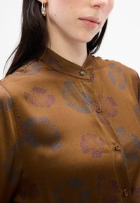 Blusa in satin marrone con motivi floreali blu e viola, colletto a punta e chiusura con bottoni e dettagli decorativi.