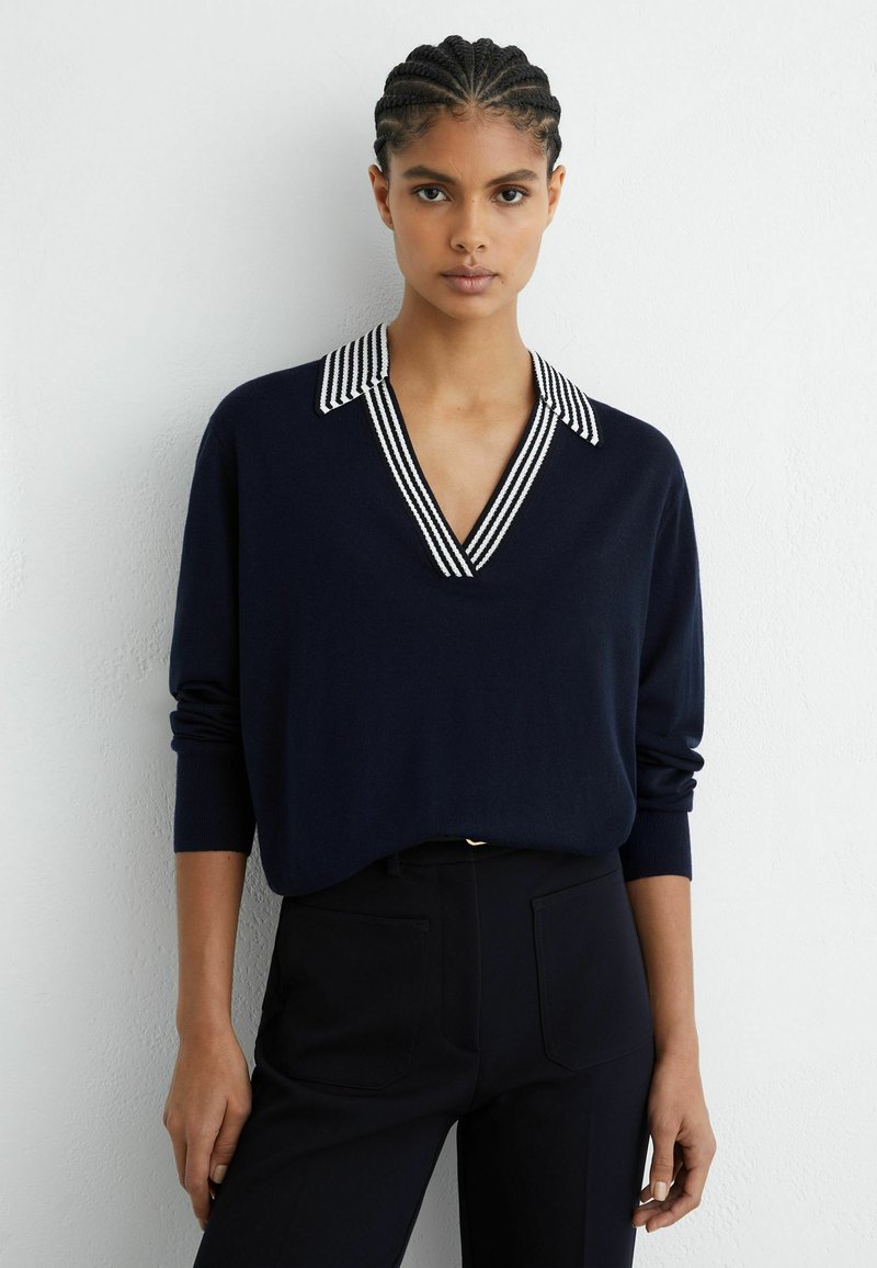 Pull-over marine à col en V avec col rayé. Manches longues, texture douce et coupe décontractée. Associé à un pantalon noir ajusté.