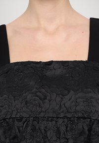 Swarte kjole med et teksturert blomsterdesign på overkroppen, med firkantet utringning og kontrastfarget glatt stoff på stroppene.