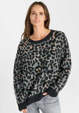 Vrouw draagt een grijs en zwart luipaardprint trui, handen gedeeltelijk in de zakken van lichtgrijze spijkerbroek, kijkt vooruit met een neutrale uitdrukking.