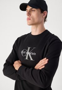 Jaunas vyras dėvi juodą Calvin Klein džemperį ir juodą beisbolo kepuraitę, susikryžiavęs rankas, žiūri į šoną prieš paprastą foną.