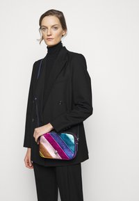 Borsa a tracolla metallica con strisce diagonali arcobaleno, dotata di chiusura a zip, superficie liscia e tracolla removibile.