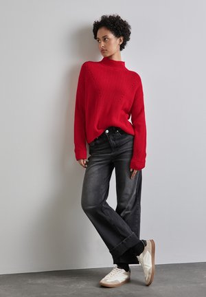 Roter gerippter Pullover mit hochgeschlossenem Kragen, kombiniert mit lockeren, dunkelgrauen Jeans. Weiße Sneakers mit beigen Akzenten. Schlichter, moderner Stil.