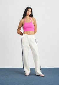 Γυναίκα με μακριά σκούρα μαλλιά στέκεται σε μπλε πάτωμα φορώντας ροζ αμάνικη κορυφή τύπου crop, λευκό υφασμάτινο παντελόνι με ναυτικές ρίγες και ροζ παπούτσια.