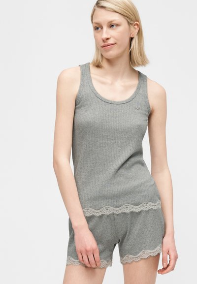 Fiatal nő szürke bordázott topban és hozzáillő rövidnadrágban, csipkés szegéllyel, világos, egyszerű háttér előtt állva.